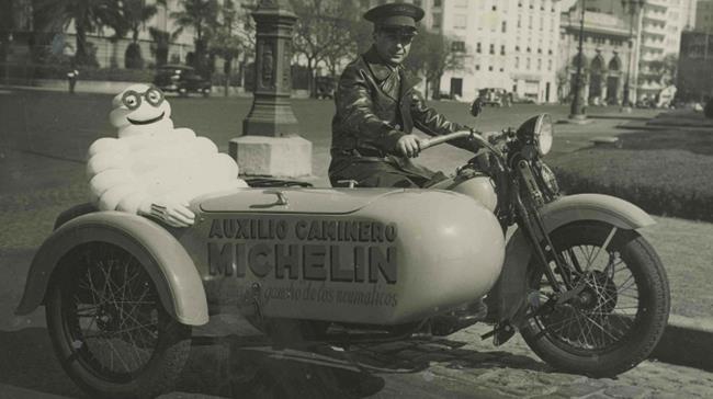 Stary motocykl, kieruje nim człowiek, a w koszu siedzi Bibendum